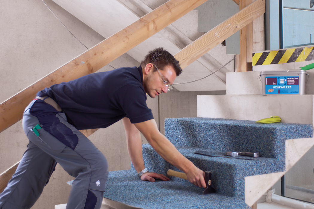 A construction worker is fitting a blue recycled carpet onto a set of unfinished concrete stairs inside a building under renovation. He is kneeling and leaning forward, wearing safety glasses, a navy polo shirt and grey work trousers, and using a rubber mallet to press the carpet firmly into place; nearby are a trowel, a tape measure and a plastic bucket labeled 'UZIN WK 222'. The scene is well lit with natural light, temporary wooden railing supports are visible, and the focus is on the careful, hands-on installation and the textured appearance of the carpet on the stair treads.