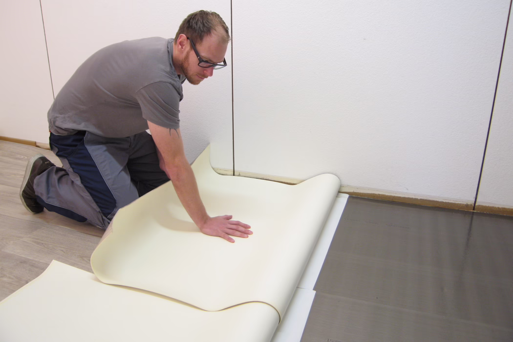 UZIN_Application Image_Sigan 2_ 2_5894_2018-10 A man wearing glasses and work clothes kneels on a light wooden subfloor as he carefully unrolls and positions a wide, cream-colored vinyl sheet against a white wall. He presses the sheet with one hand to smooth it while aligning it over a dark underlayment; nearby are visible seams and a strip of exposed subfloor along the wall. The close-up scene documents a floor installation process in a simple, well-lit room, emphasizing the craftsman's deliberate movements, the textures of the materials and the contrast between the pale vinyl and the darker underlayer.